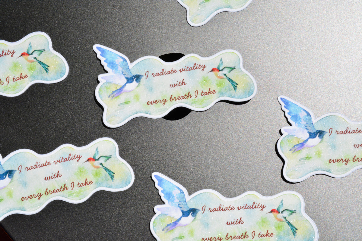 Several die-cut sticker shapes are scattered on a dark gray surface. Each sticker features a watercolor-style blue bird with green and red accents, plus the printed phrase 'I radiate vitality with every breath I take' in brown text.