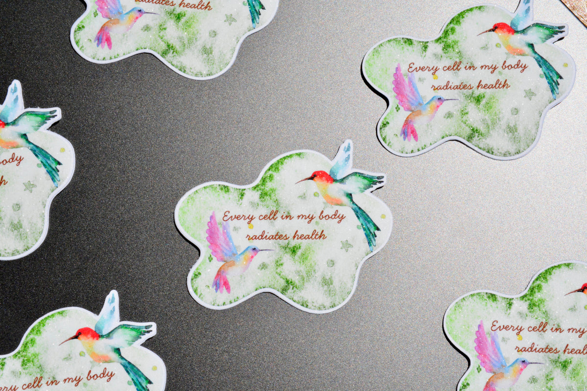Several cloud-shaped stickers with watercolor hummingbirds and the phrase 'Every cell in my body radiates health' are arranged on a dark gray surface.