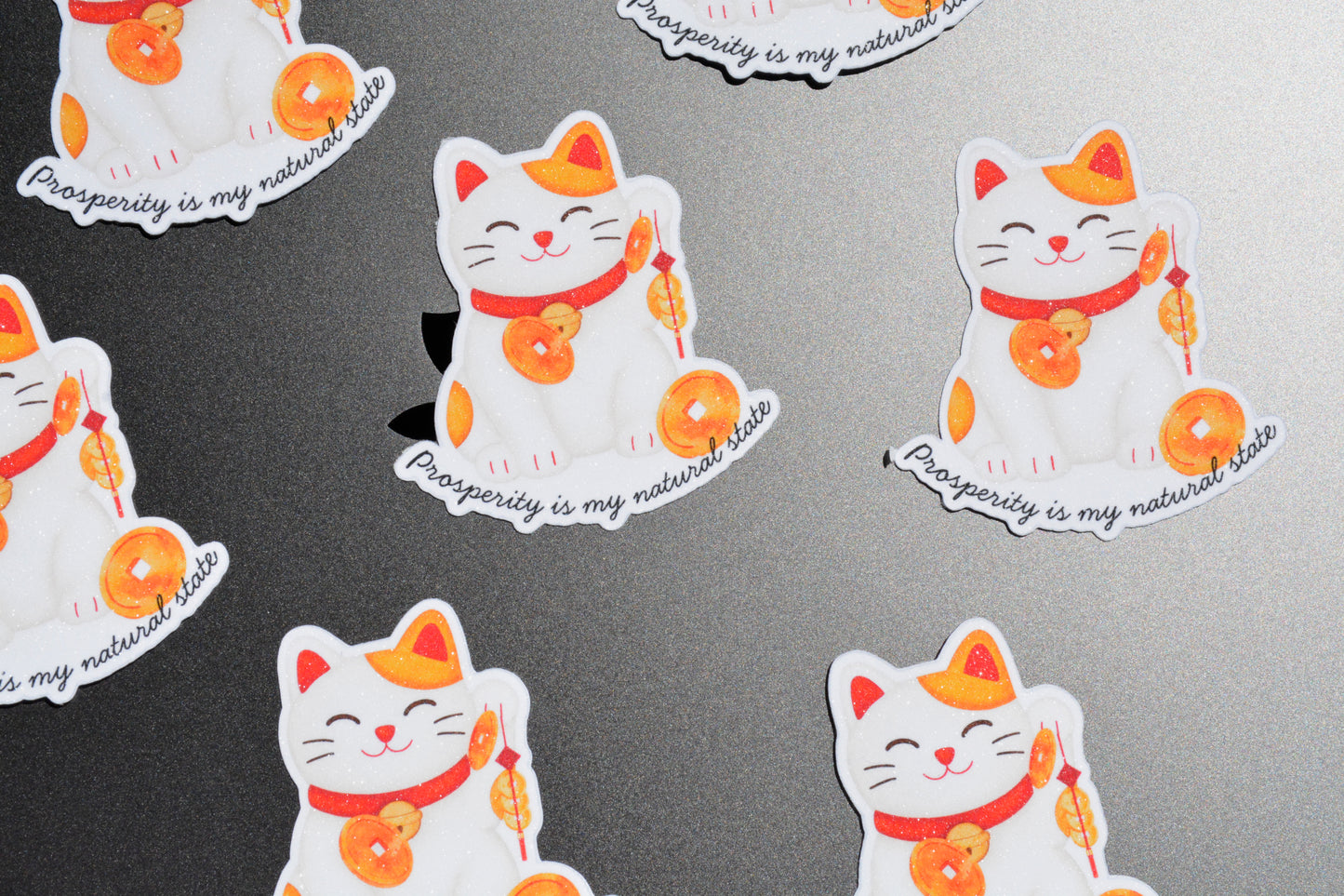 White and orange lucky cat stickers with red accents and the phrase 'Prosperity is my natural state' printed in script, arranged on a dark gray textured surface.