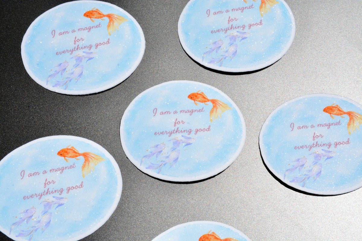 Five round coasters with a light blue watercolor background, featuring an orange goldfish, purple flowers, and the text 'I am a magnet for everything good' on a dark gray surface.