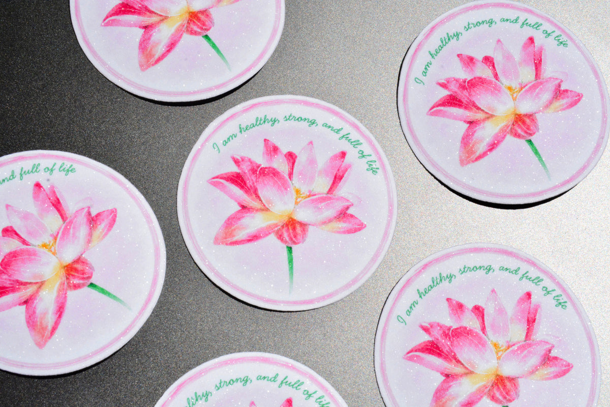 A set of round coasters featuring a pink watercolor lotus flower and the quote 'I am healthy, strong, and full of life' in green script, arranged on a gray textured surface.
