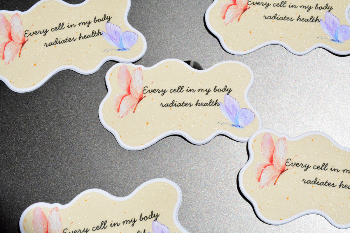 Several die-cut sticker tags with a wavy cloud shape are arranged on a gray textured surface. Each tag has a cream background with a small watercolor pink flower, a lavender butterfly, and the printed phrase 'Every cell in my body radiates health' in black script.