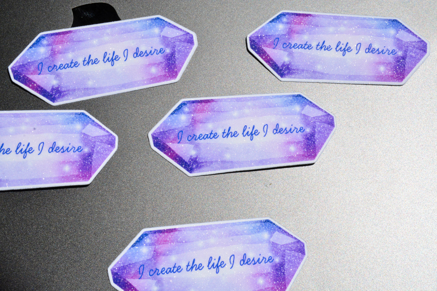 Five identical die-cut sticker labels with a purple and blue galaxy gemstone design, each printed with the phrase 'I create the life I desire' in blue script, arranged on a gray surface.