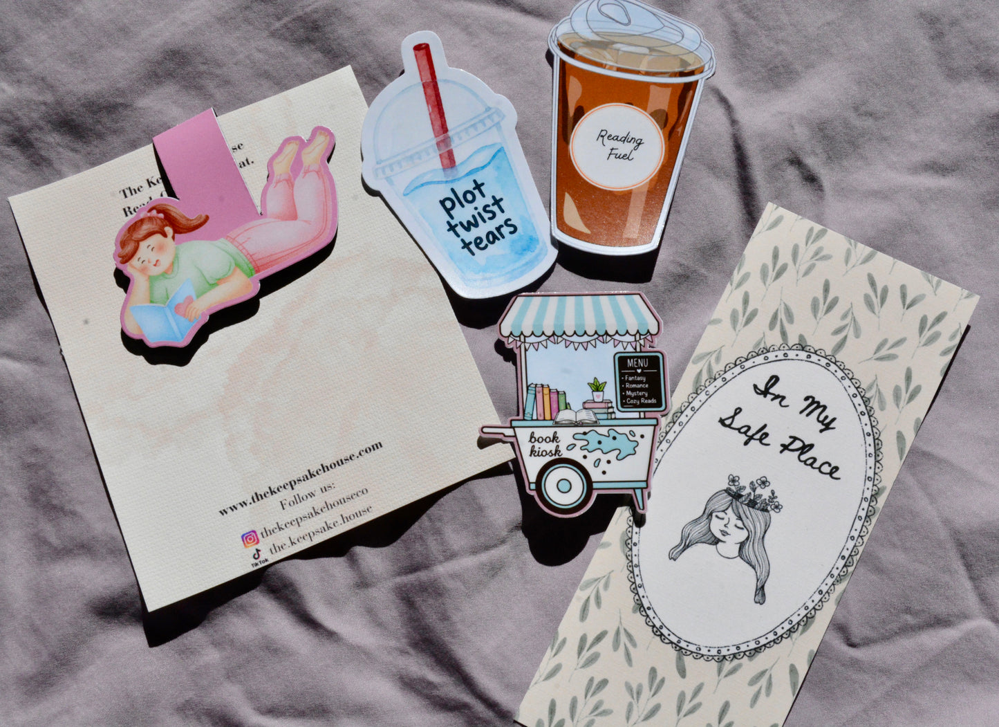A flat-lay of several printed paper items on a gray fabric background, including a cream card with a pink illustrated character, a blue drink cup sticker reading 'plot twist teas,' a brown coffee cup sticker, a small food cart sticker, and a cream oval pin or patch with the text 'In My Left Place' and a black-haired illustrated woman.