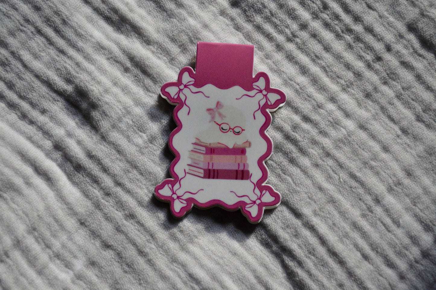 A small pink and white decorative accessory with a scalloped edge with stacked books and sleeping ghost, placed on a textured gray fabric background.