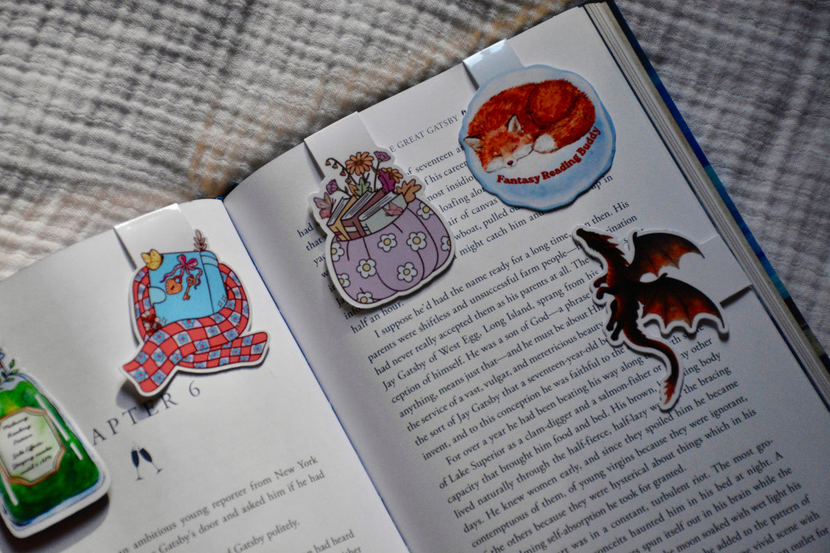 A set of whimsical die-cut magnetic bookmarks shown attached to an open book. The bookmarks include a green potion bottle, a purple owl, a red cat in a blue hat, and a black dragon, resting on the pages next to printed text.