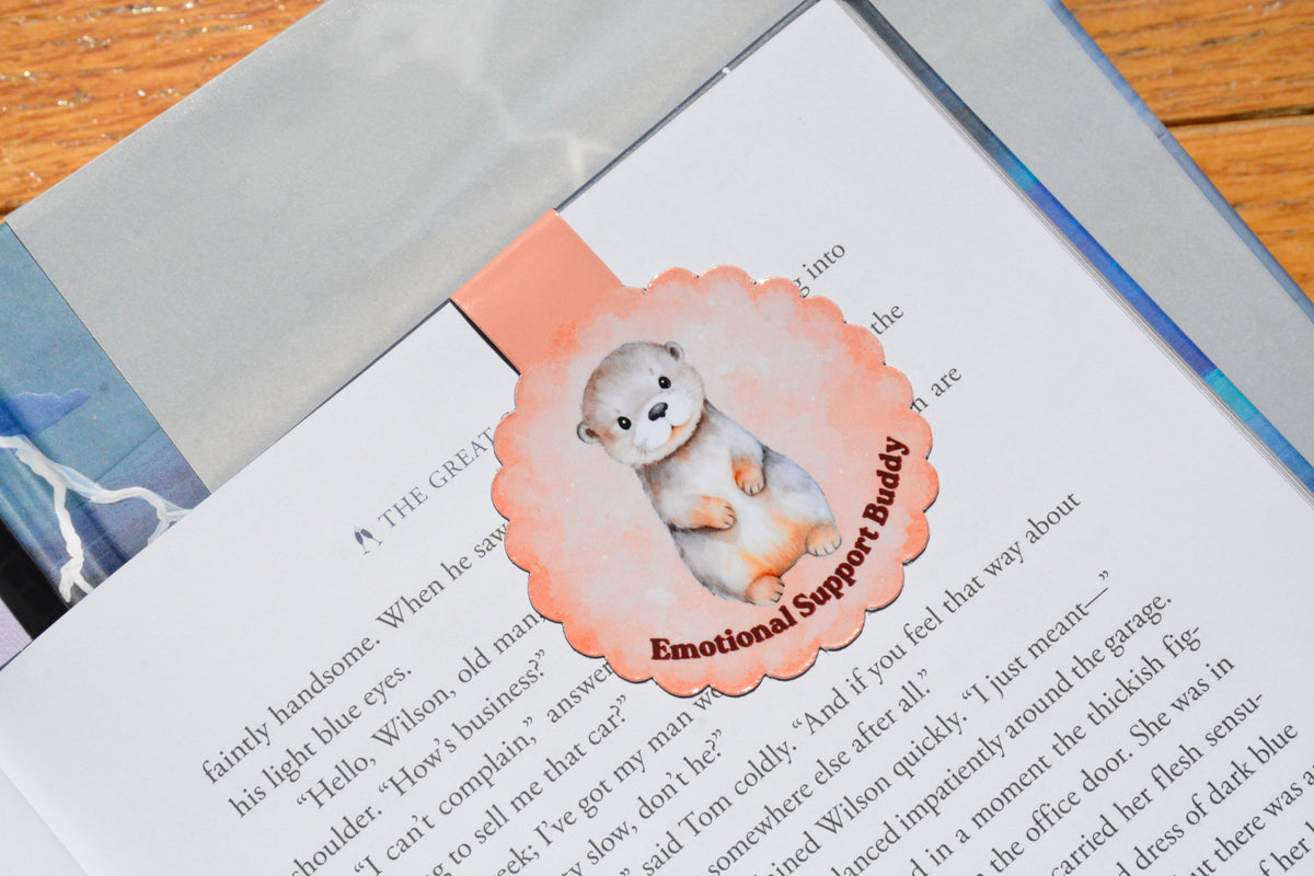 Handmade magnetic otter bookmark with a soft peach watercolor design, shown marking a page in a novel, perfect for readers, students, and book lovers.