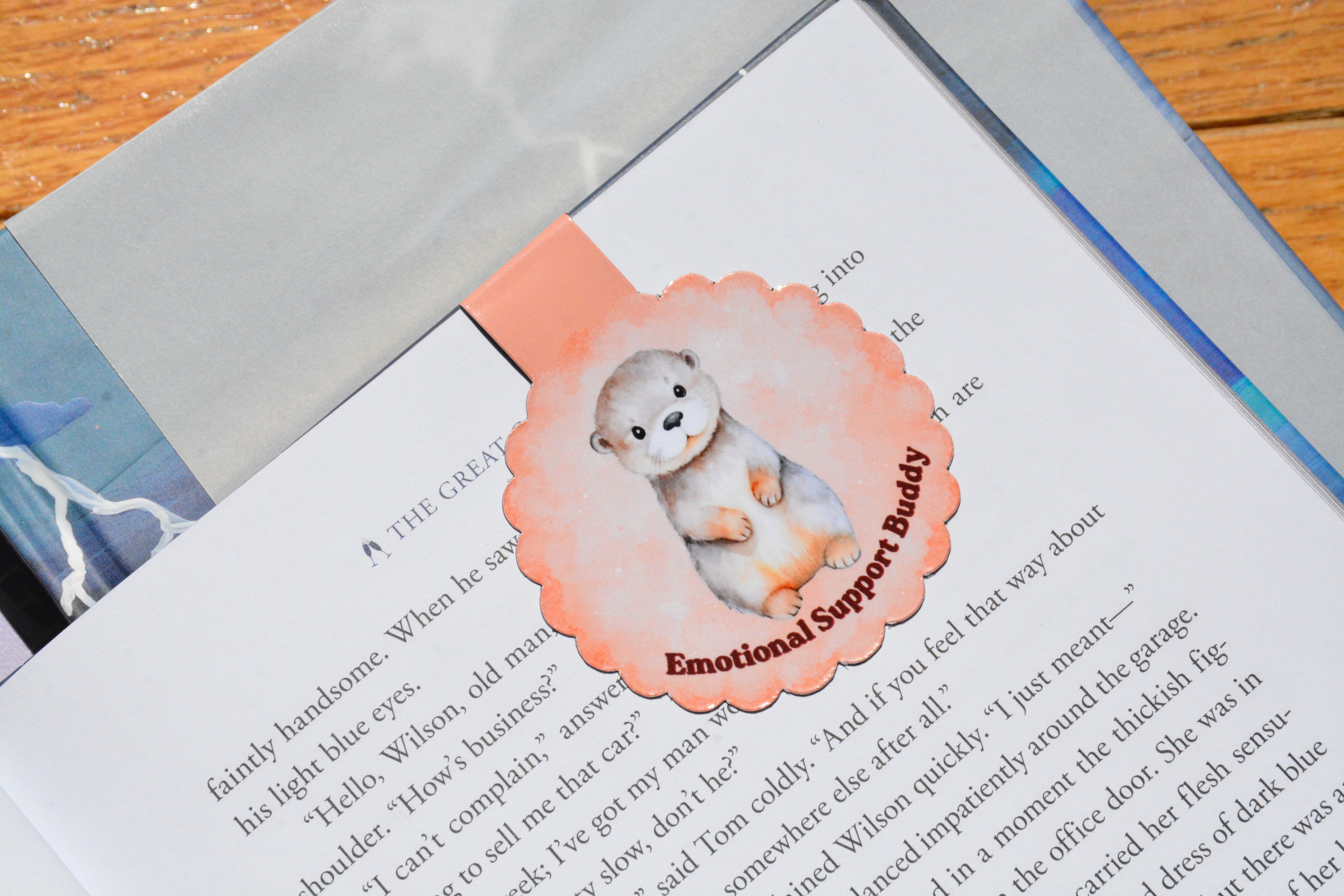Handmade magnetic otter bookmark with a soft peach watercolor design, shown marking a page in a novel, perfect for readers, students, and book lovers.