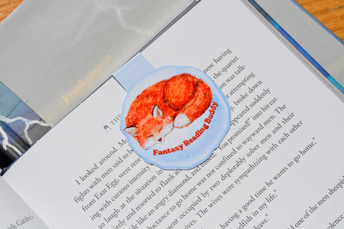 Handmade magnetic fox bookmark with a cozy watercolor sleeping fox illustration marking a page in a novel, perfect for fantasy readers and book lovers.
