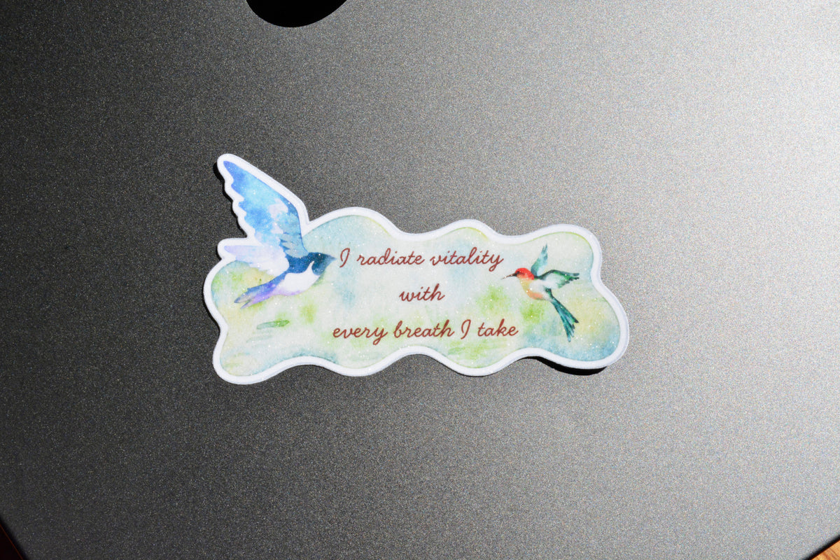 A die-cut sticker featuring a watercolor design of two birds and the text 'I radiate vitality with every breath I take', placed on a dark gray metallic surface.