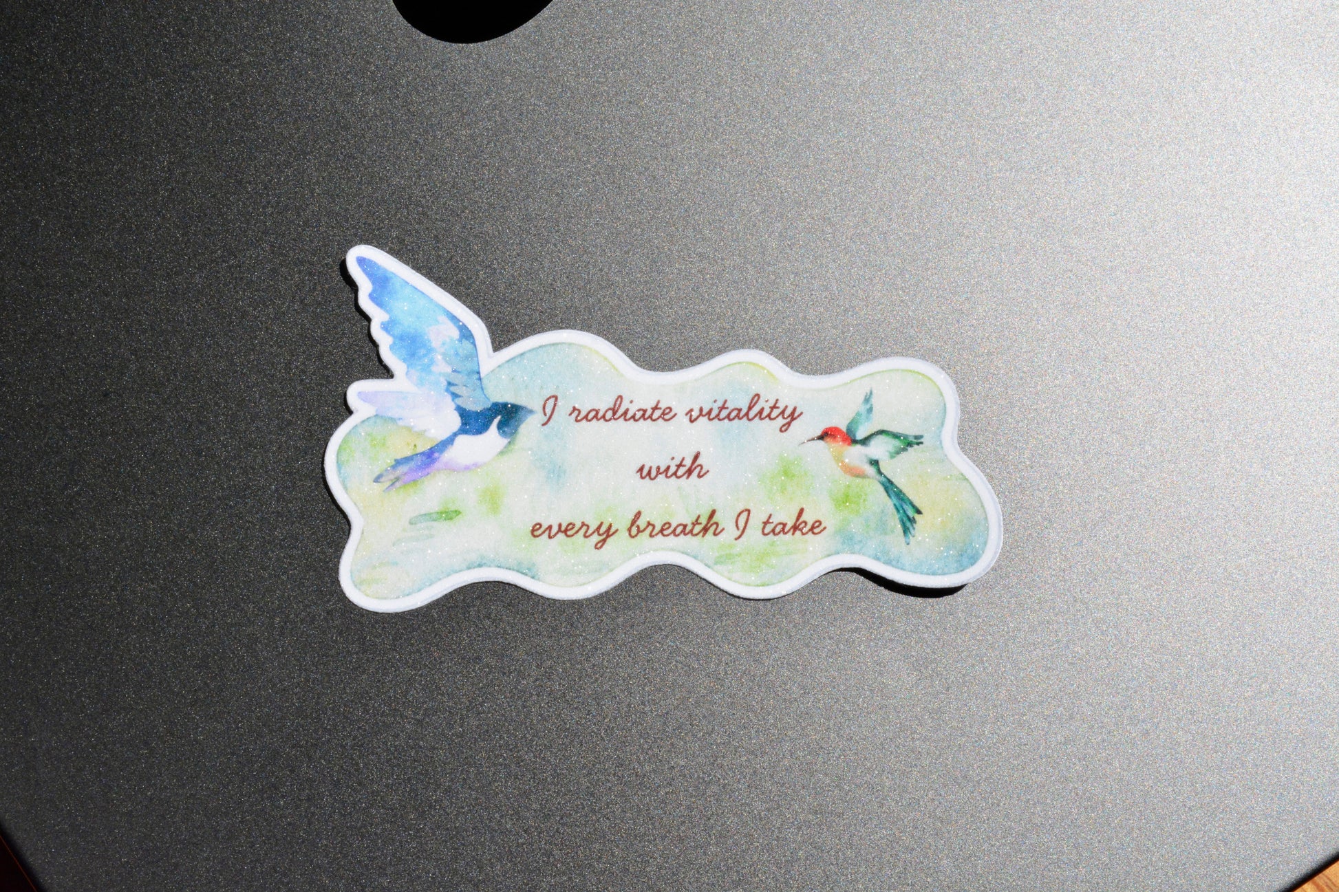 A die-cut sticker featuring a watercolor design of two birds and the text 'I radiate vitality with every breath I take', placed on a dark gray metallic surface.