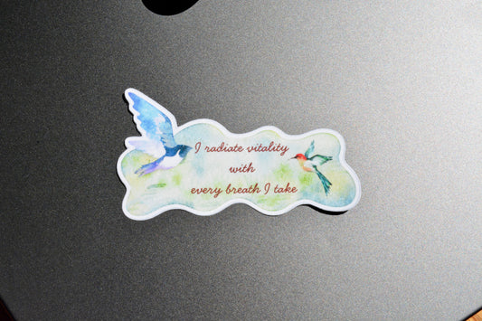 A die-cut sticker featuring a watercolor design of two birds and the text 'I radiate vitality with every breath I take', placed on a dark gray metallic surface.