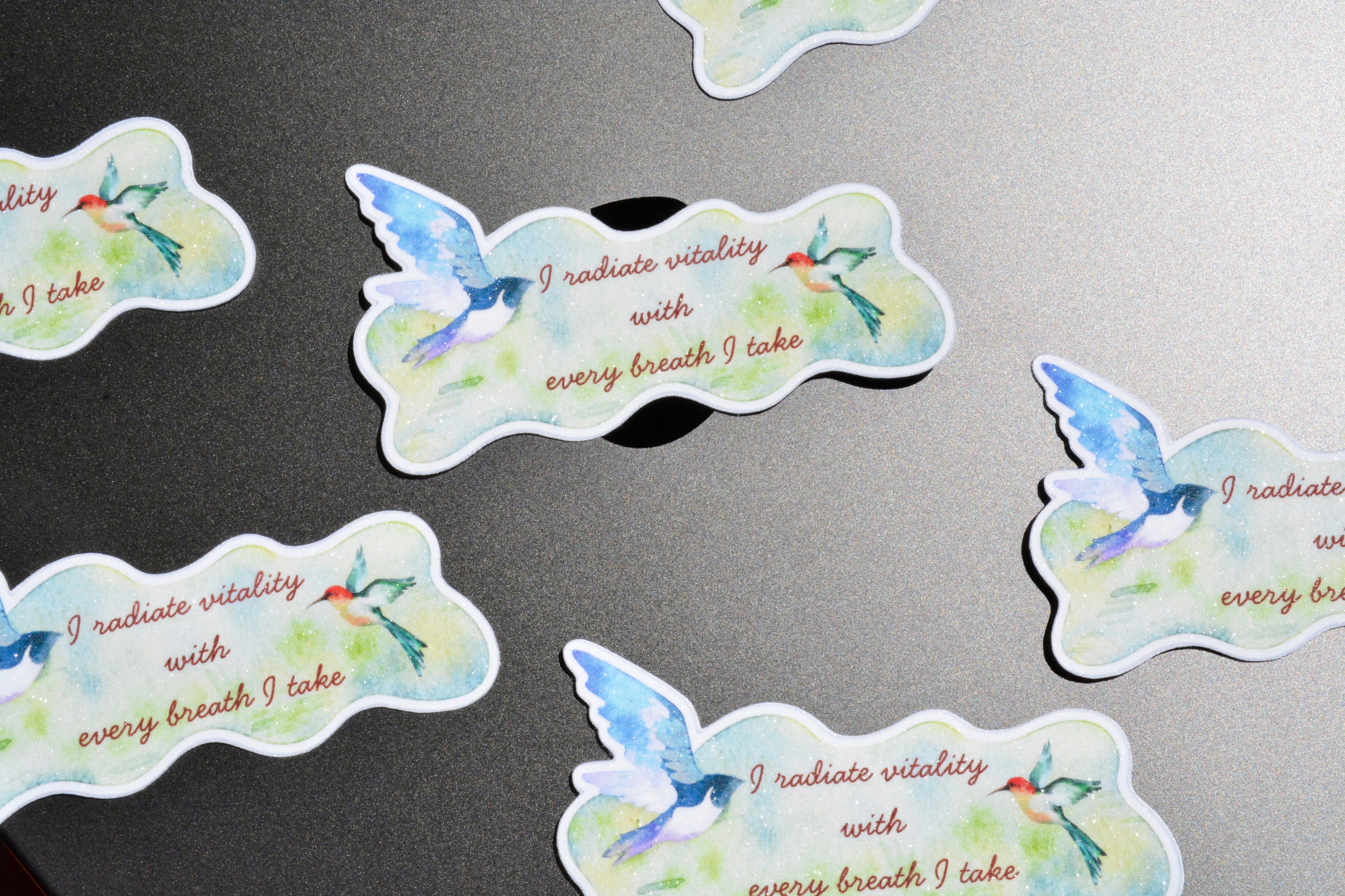 Several die-cut sticker shapes are scattered on a dark gray surface. Each sticker features a watercolor-style blue bird with green and red accents, plus the printed phrase 'I radiate vitality with every breath I take' in brown text.