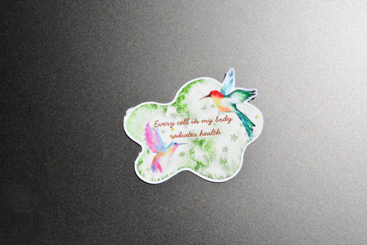 A die-cut sticker featuring two colorful hummingbirds and the printed phrase 'Every cell in my body indicates health' on a white background with green watercolor splashes, shown on a dark gray textured surface.
