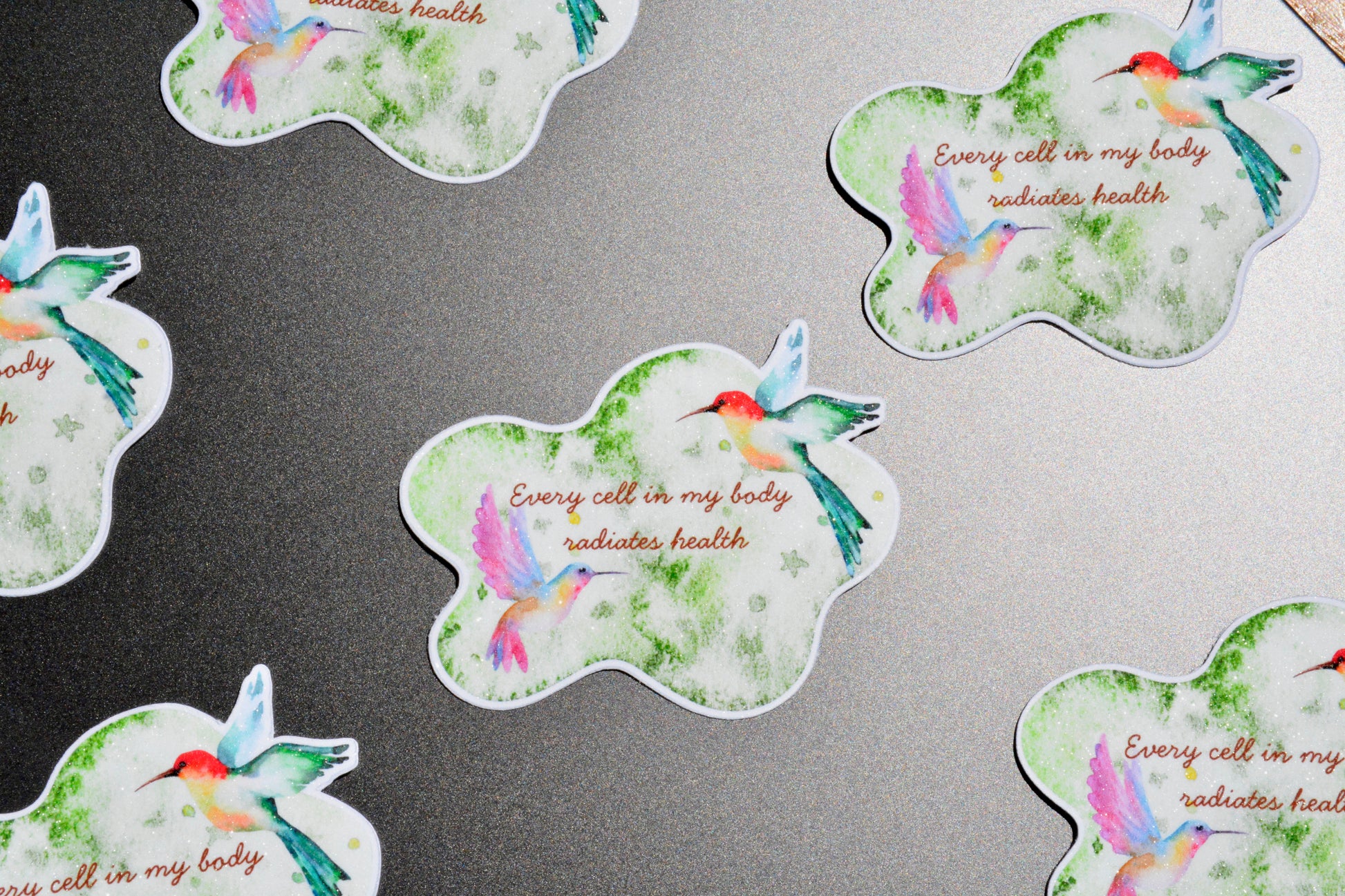 Several cloud-shaped stickers with watercolor hummingbirds and the phrase 'Every cell in my body radiates health' are arranged on a dark gray surface.