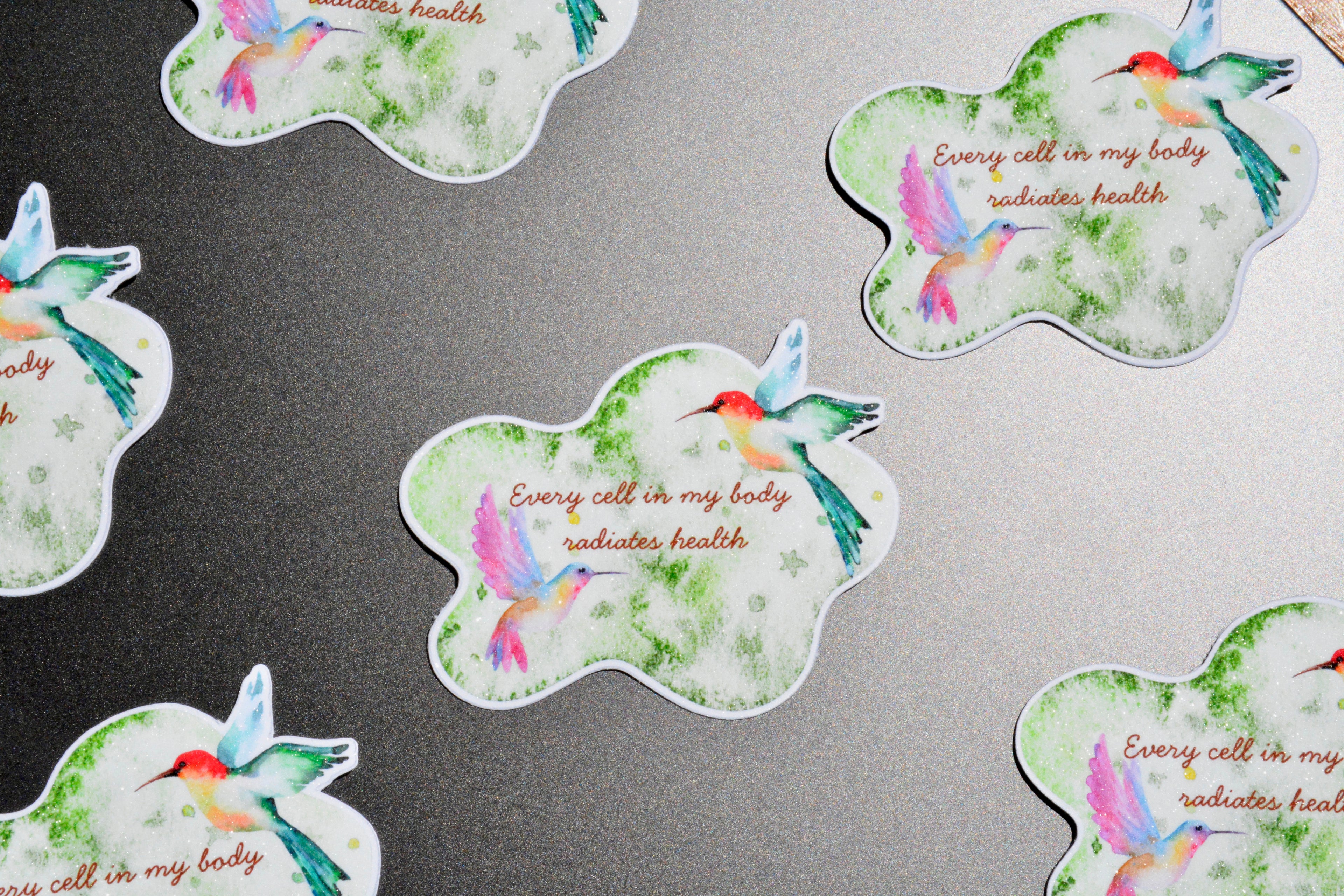 Several cloud-shaped stickers with watercolor hummingbirds and the phrase 'Every cell in my body radiates health' are arranged on a dark gray surface.