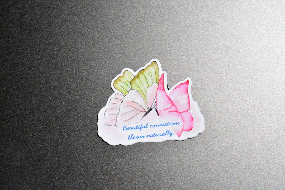 A die-cut sticker featuring three watercolor-style butterflies in pink, white, and light green, with the blue script text 'Beautiful connections bloom naturally' on a white background, placed on a dark gray textured surface.