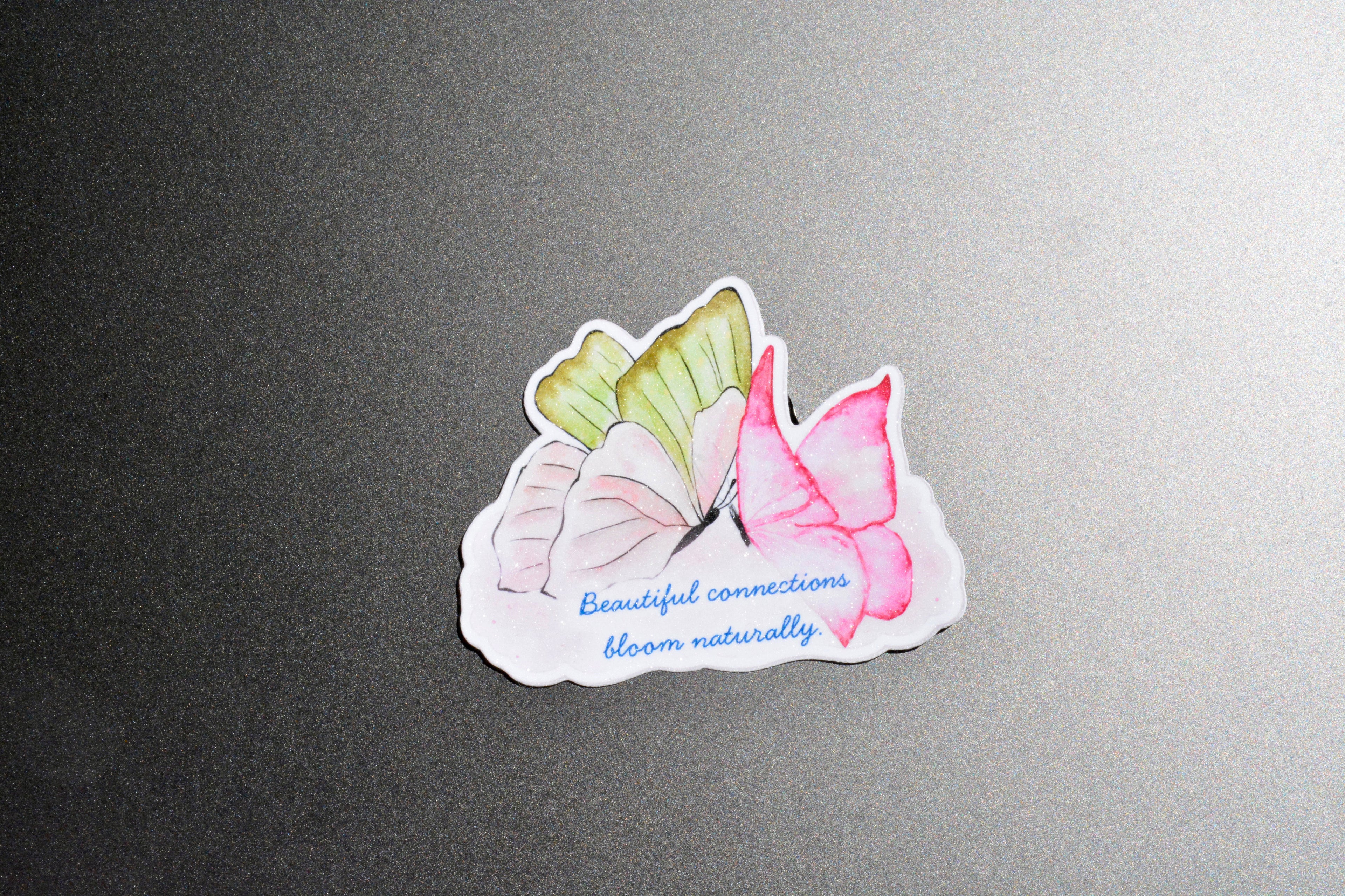 A die-cut sticker featuring three watercolor-style butterflies in pink, white, and light green, with the blue script text 'Beautiful connections bloom naturally' on a white background, placed on a dark gray textured surface.