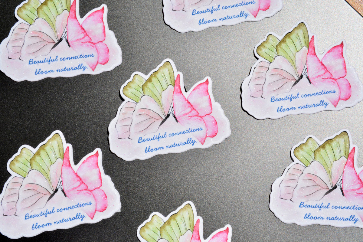 Several die-cut stickers featuring watercolor butterflies in pink, green, and white with the text 'Beautiful connections bloom naturally' in blue, arranged on a dark gray textured surface.
