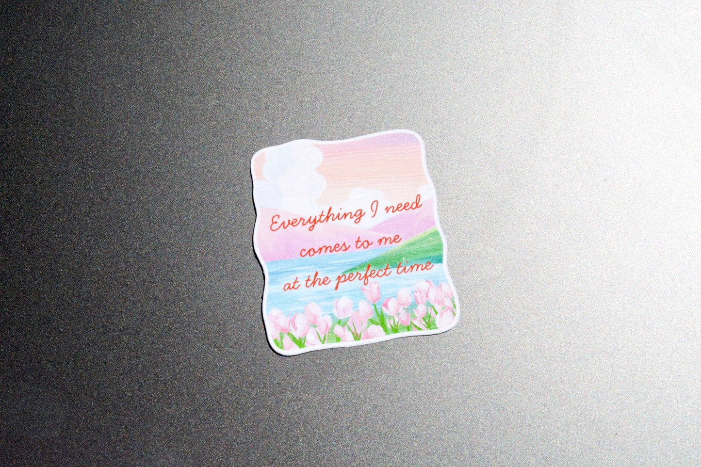 A die-cut sticker with a pastel landscape design featuring pink clouds, blue sky, green grass, and pink tulips, with the text 'Everything I need comes to me at the perfect time' in orange script, shown on a dark gray textured surface.