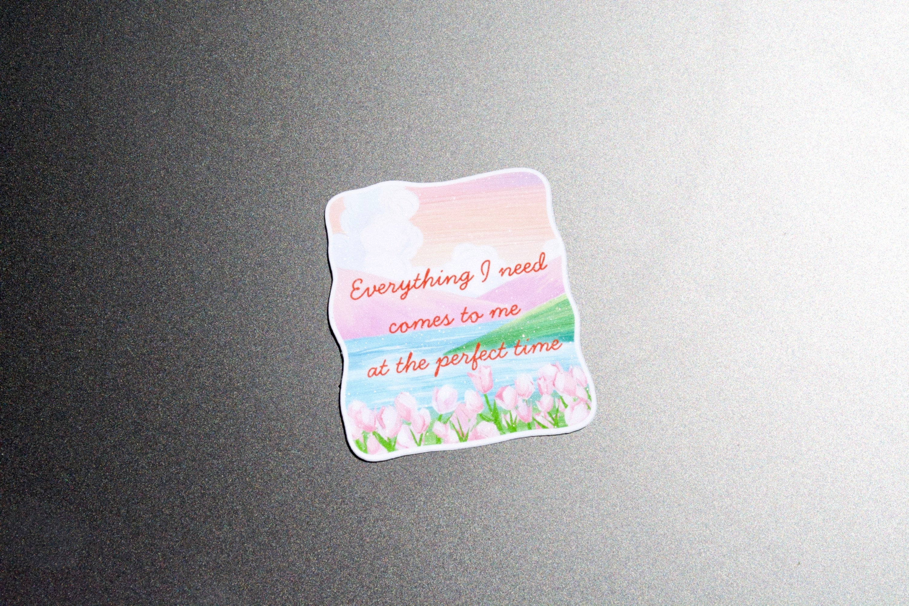 A die-cut sticker with a pastel landscape design featuring pink clouds, blue sky, green grass, and pink tulips, with the text 'Everything I need comes to me at the perfect time' in orange script, shown on a dark gray textured surface.