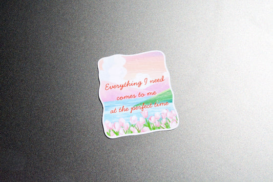 A die-cut sticker with a pastel landscape design featuring pink clouds, blue sky, green grass, and pink tulips, with the text 'Everything I need comes to me at the perfect time' in orange script, shown on a dark gray textured surface.
