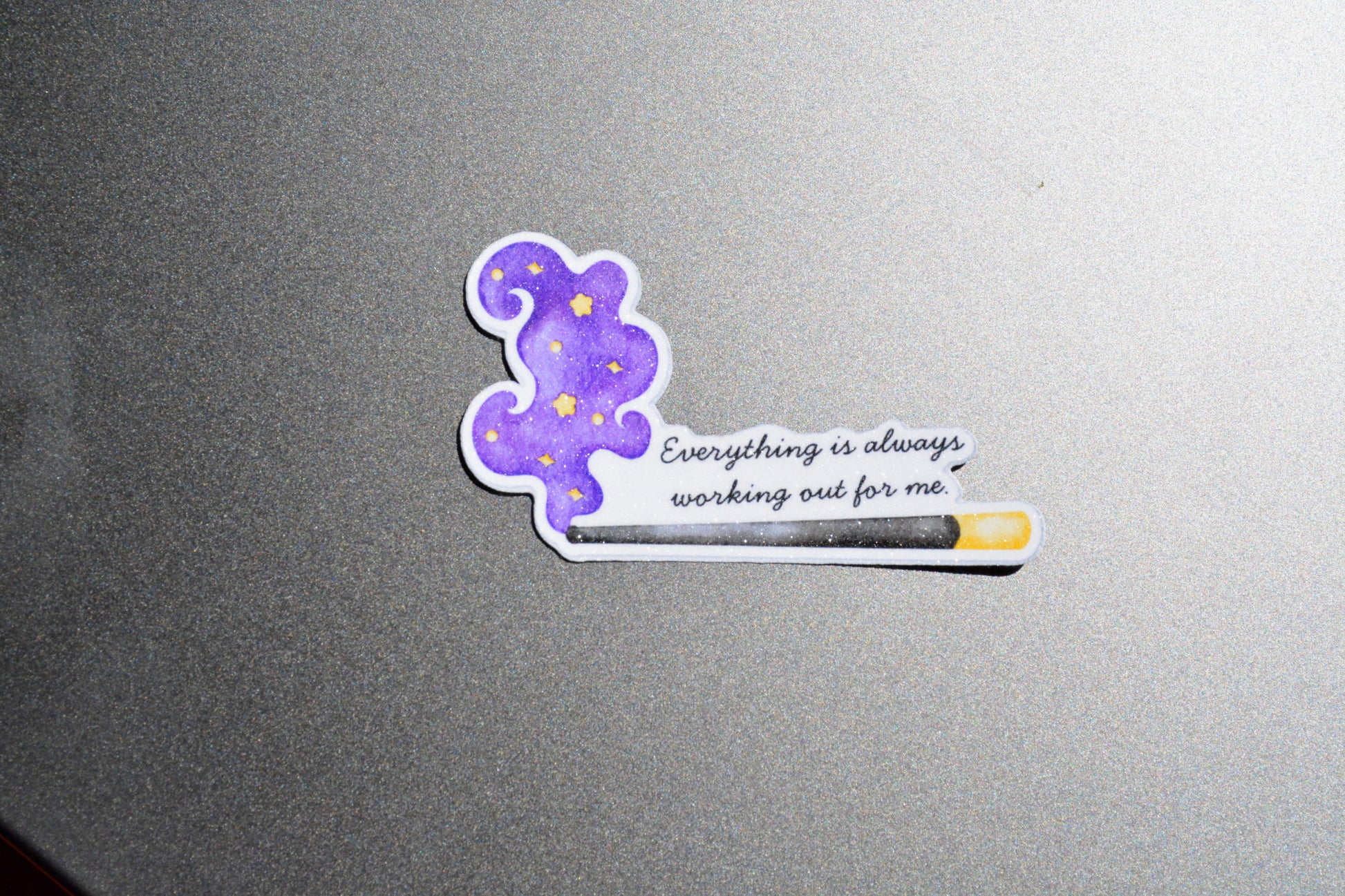 A die-cut sticker featuring a purple cloud with yellow stars and the quote 'Everything is always working out for me' next to a progress bar, shown on a textured gray surface.