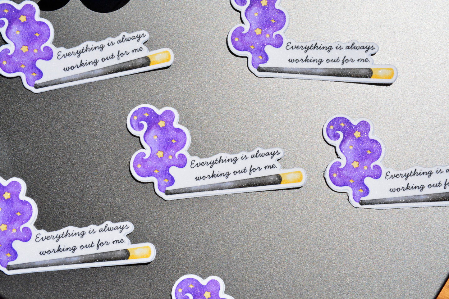 Several die-cut stickers featuring a purple wizard hat and wand design with the text 'Everything is always working out for me' are arranged on a dark gray surface.