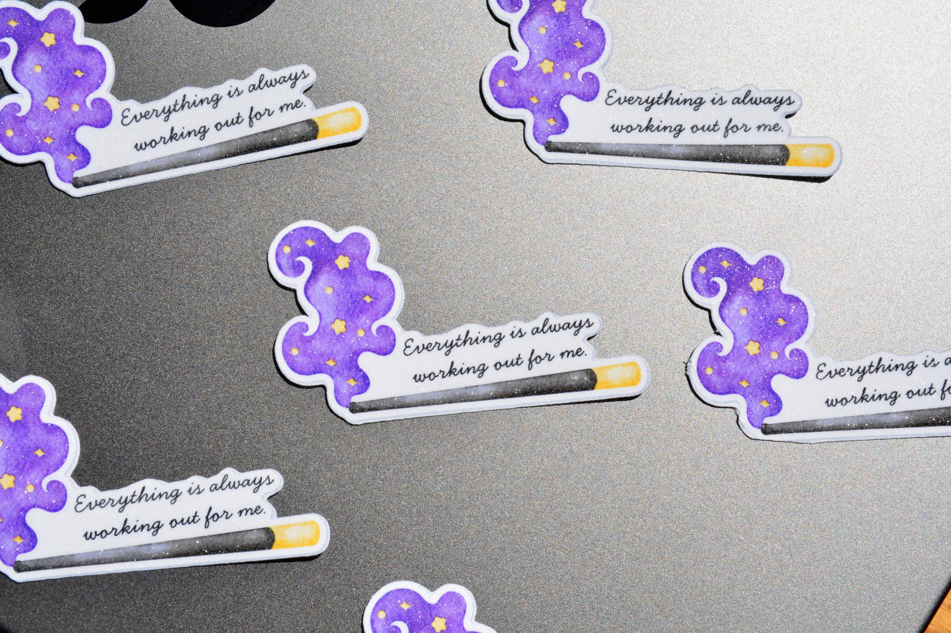Several die-cut stickers featuring a purple wizard hat and wand design with the text 'Everything is always working out for me' are arranged on a dark gray surface.
