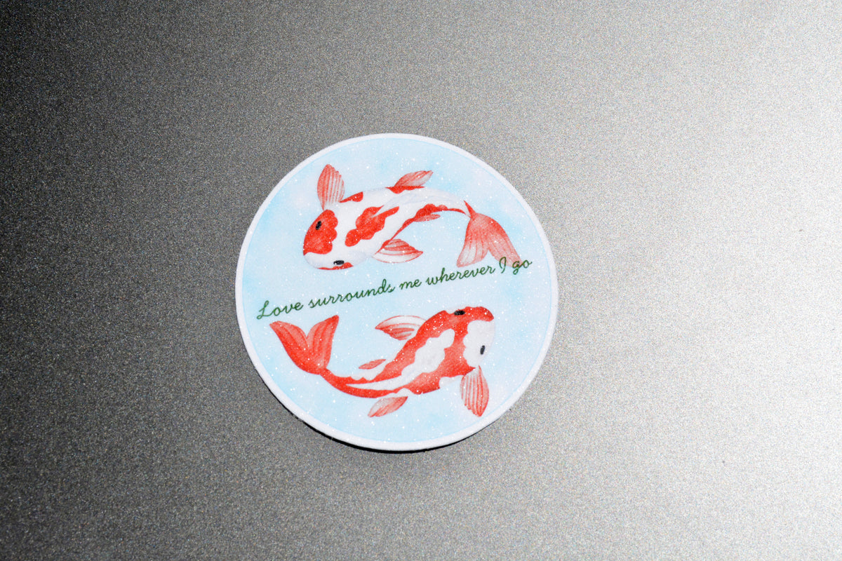 A round sticker featuring two red and white koi fish on a light blue background, with the green cursive text 'Love surrounds me wherever I go', displayed on a textured gray surface.