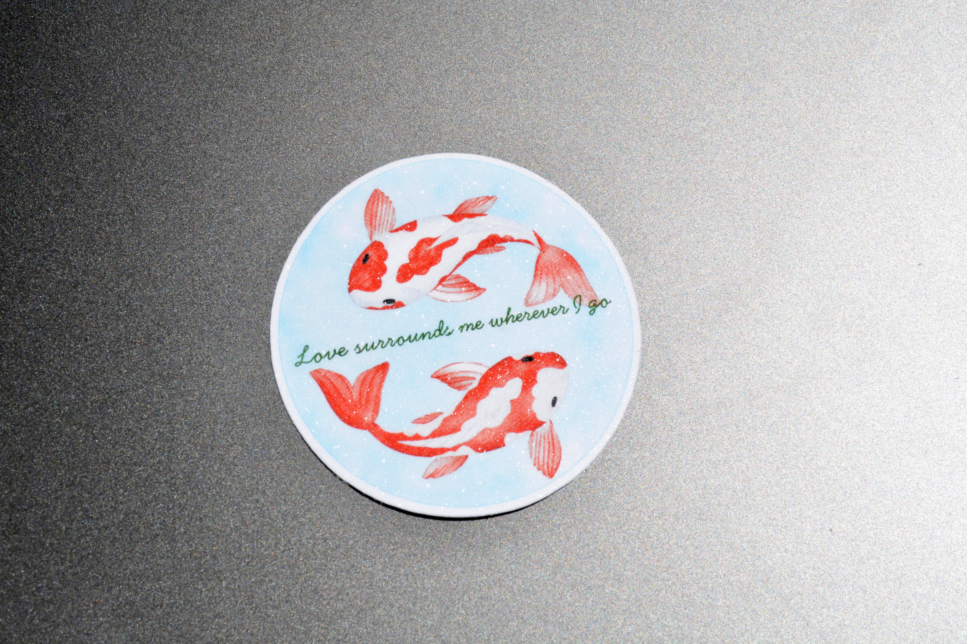 A round sticker featuring two red and white koi fish on a light blue background, with the green cursive text 'Love surrounds me wherever I go', displayed on a textured gray surface.
