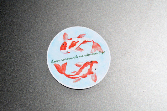A round sticker featuring two red and white koi fish on a light blue background, with the green cursive text 'Love surrounds me wherever I go', displayed on a textured gray surface.