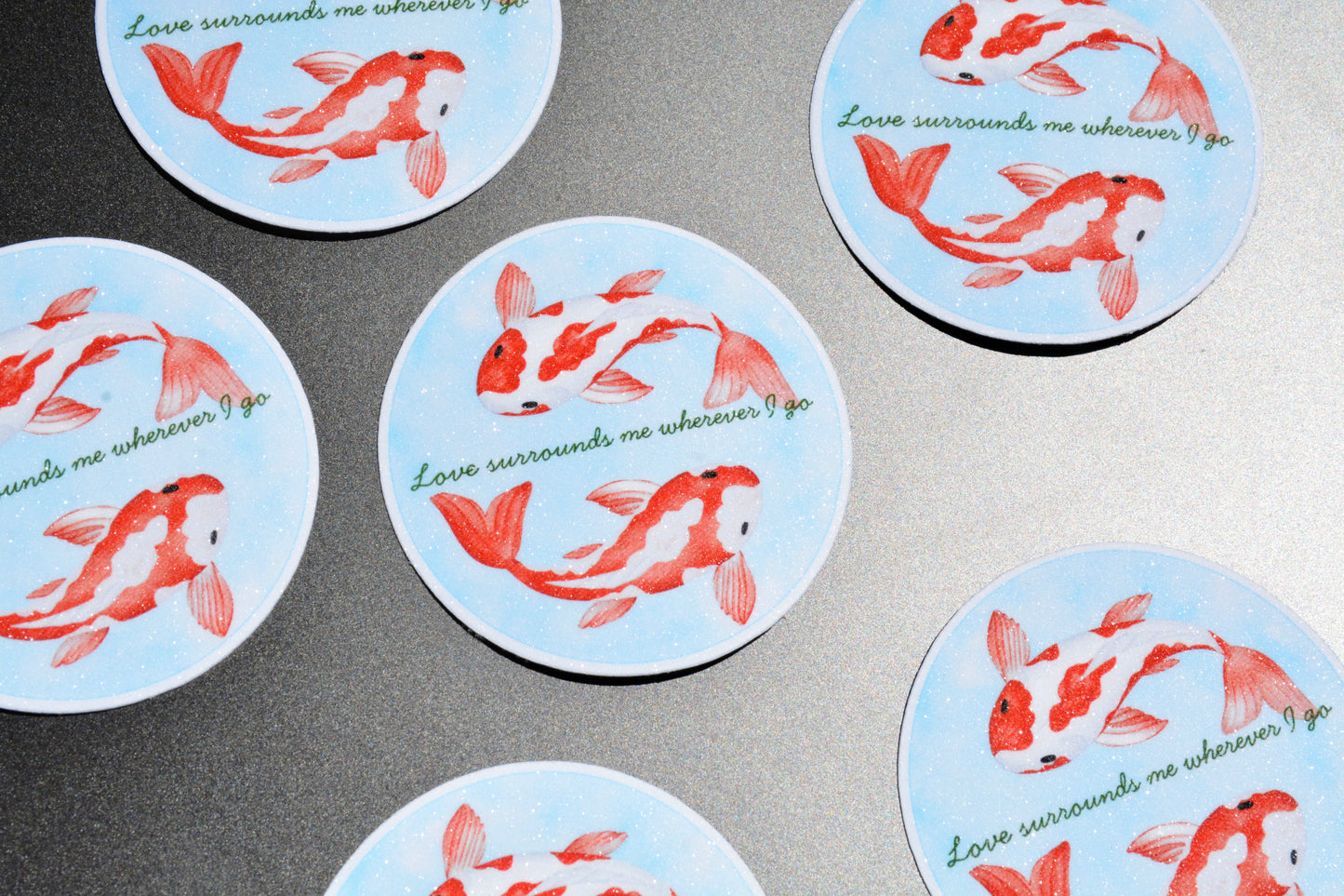 Several round coasters with a light blue background, printed with red koi fish and the phrase 'Love surrounds me wherever I go' in green script, arranged on a gray surface.