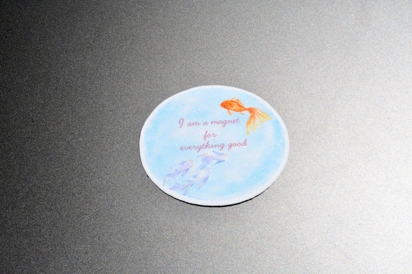 A round white coaster with a light blue watercolor background, featuring an orange goldfish, purple fish, and the text 'I am a magnet for everything good' in pink script, shown on a dark gray textured surface.