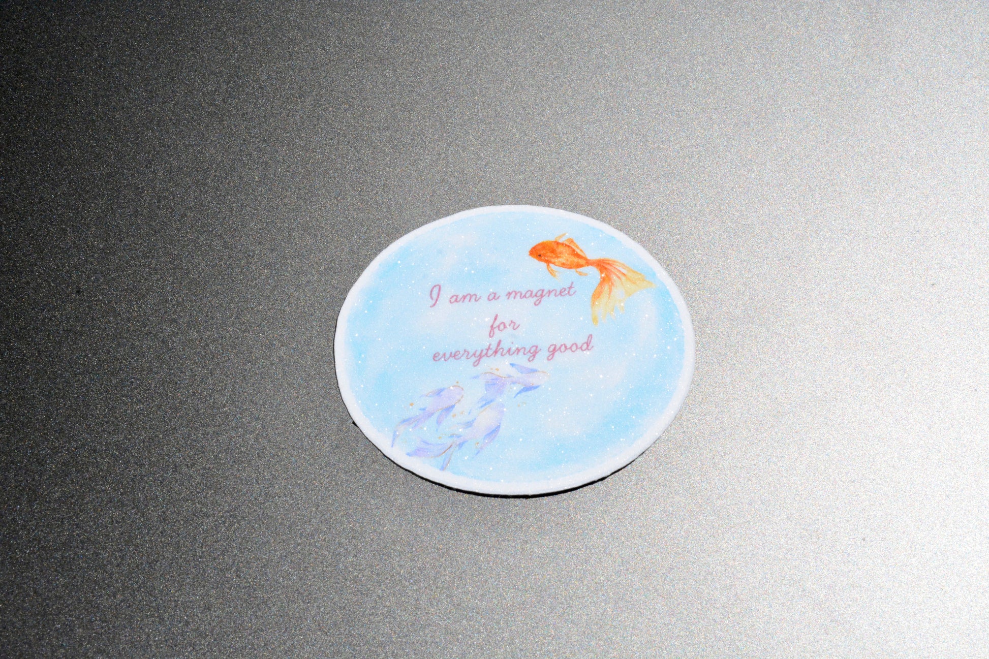 A round white coaster with a light blue watercolor background, featuring an orange goldfish, purple fish, and the text 'I am a magnet for everything good' in pink script, shown on a dark gray textured surface.