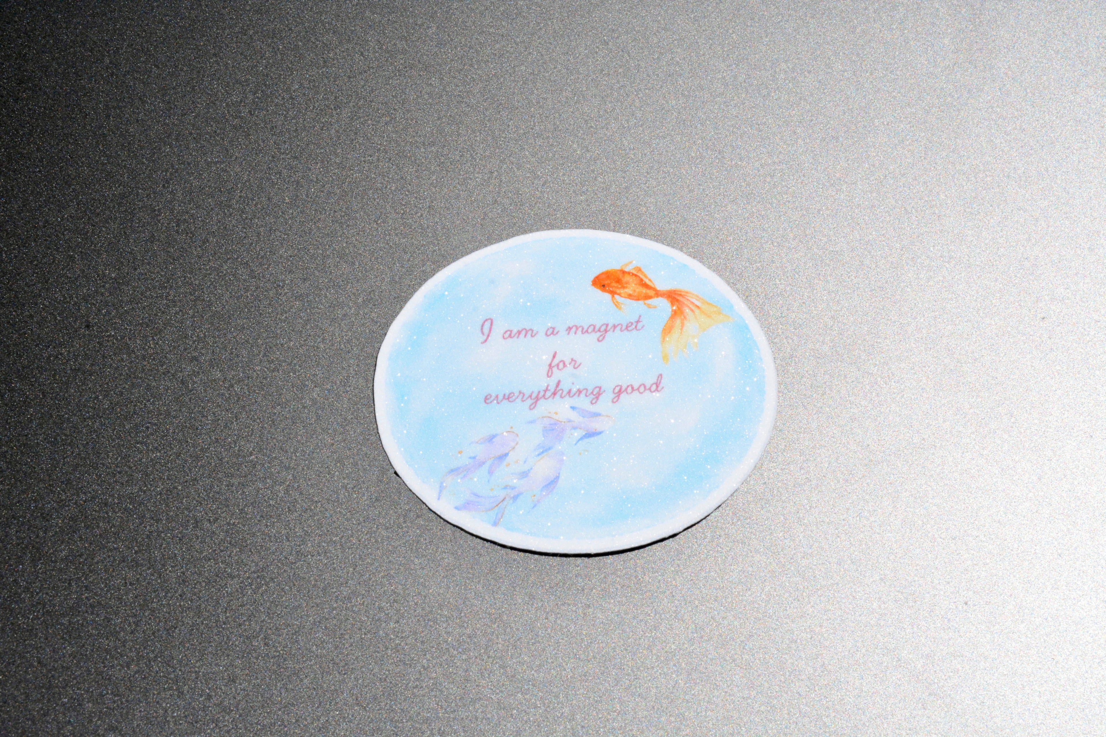 A round white coaster with a light blue watercolor background, featuring an orange goldfish, purple fish, and the text 'I am a magnet for everything good' in pink script, shown on a dark gray textured surface.