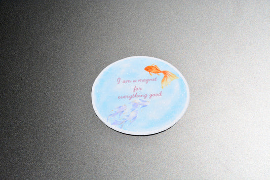 A round white coaster with a light blue watercolor background, featuring an orange goldfish, purple fish, and the text 'I am a magnet for everything good' in pink script, shown on a dark gray textured surface.