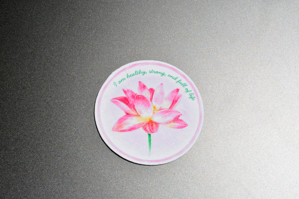 A round sticker featuring a pink watercolor lotus flower with green text that reads 'I am healthy, strong, and full of life', displayed on a textured gray background.