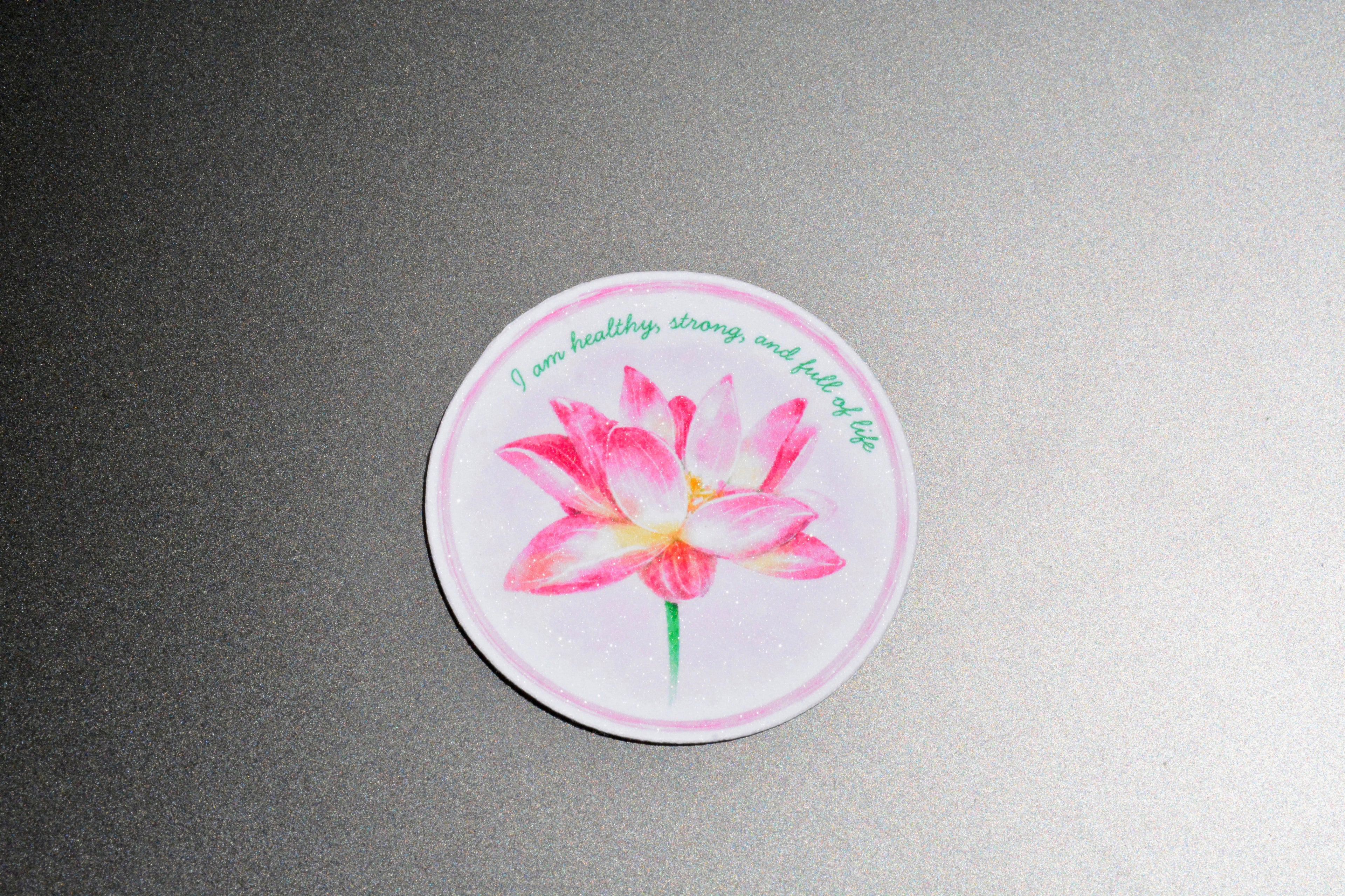 A round sticker featuring a pink watercolor lotus flower with green text that reads 'I am healthy, strong, and full of life', displayed on a textured gray background.