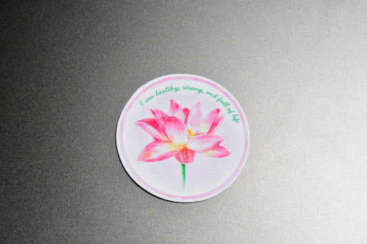 A round sticker featuring a pink watercolor lotus flower with green text that reads 'I am healthy, strong, and full of life', displayed on a textured gray background.