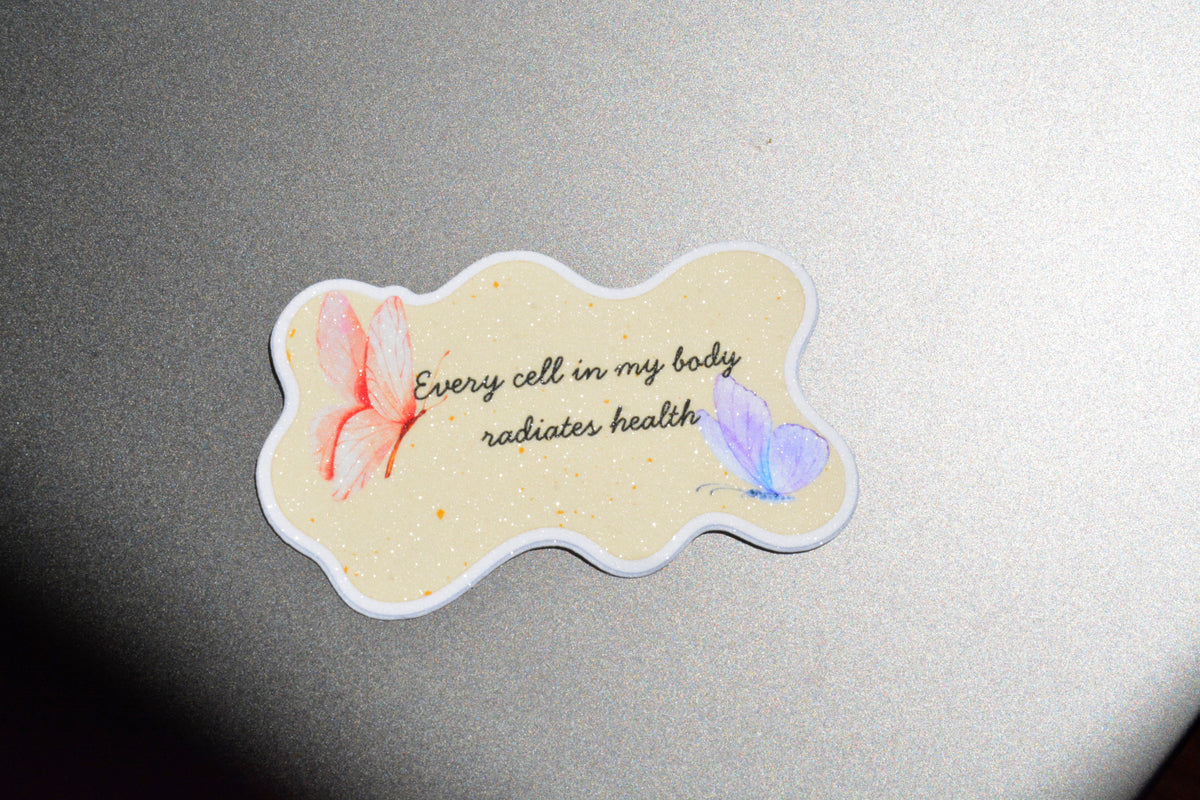 A die-cut sticker with a wavy outline featuring the text 'Every cell in my body radiates health' in black script, decorated with a pink butterfly on the left and a light purple butterfly on the right, set against a textured gray background.