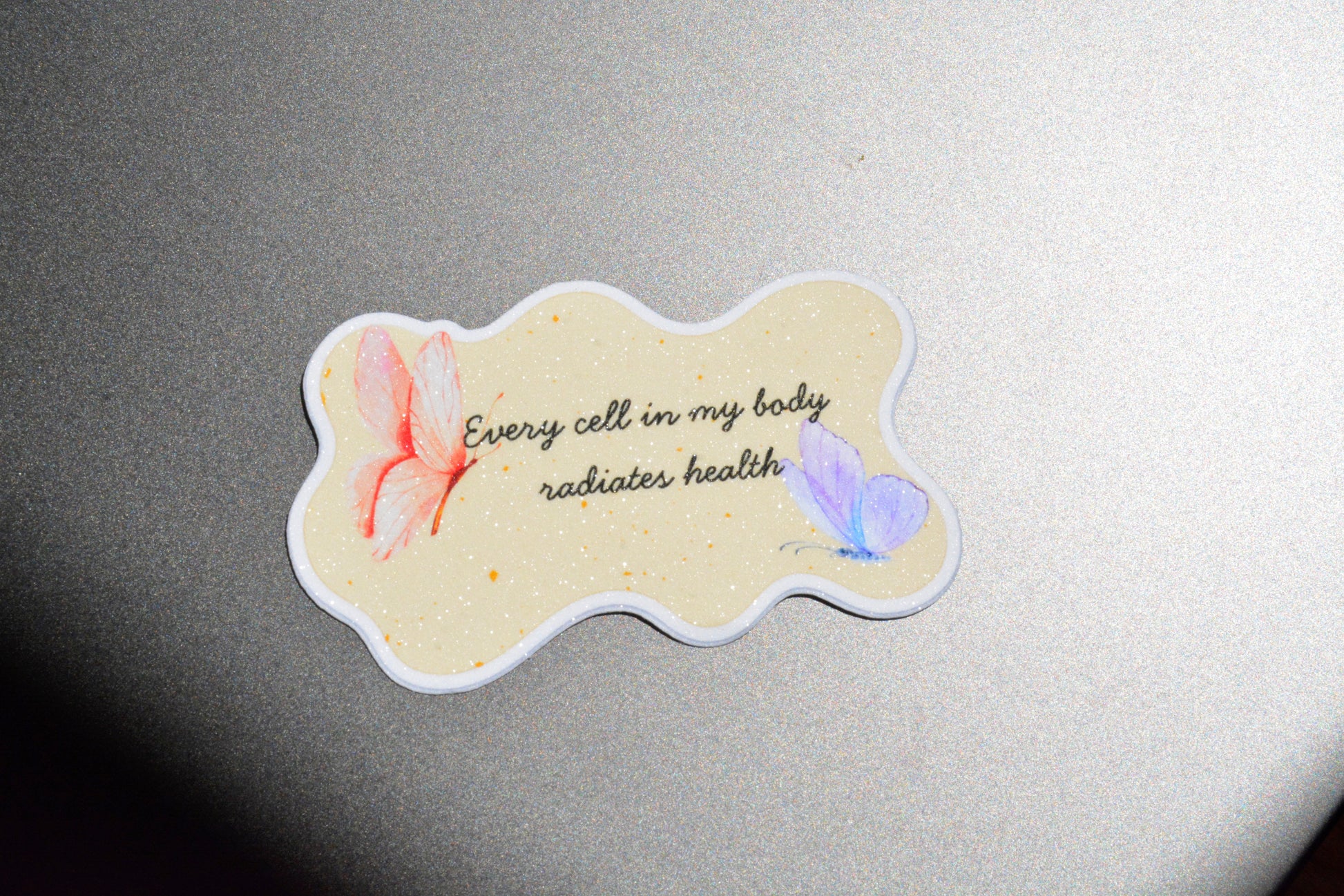 A die-cut sticker with a wavy outline featuring the text 'Every cell in my body radiates health' in black script, decorated with a pink butterfly on the left and a light purple butterfly on the right, set against a textured gray background.
