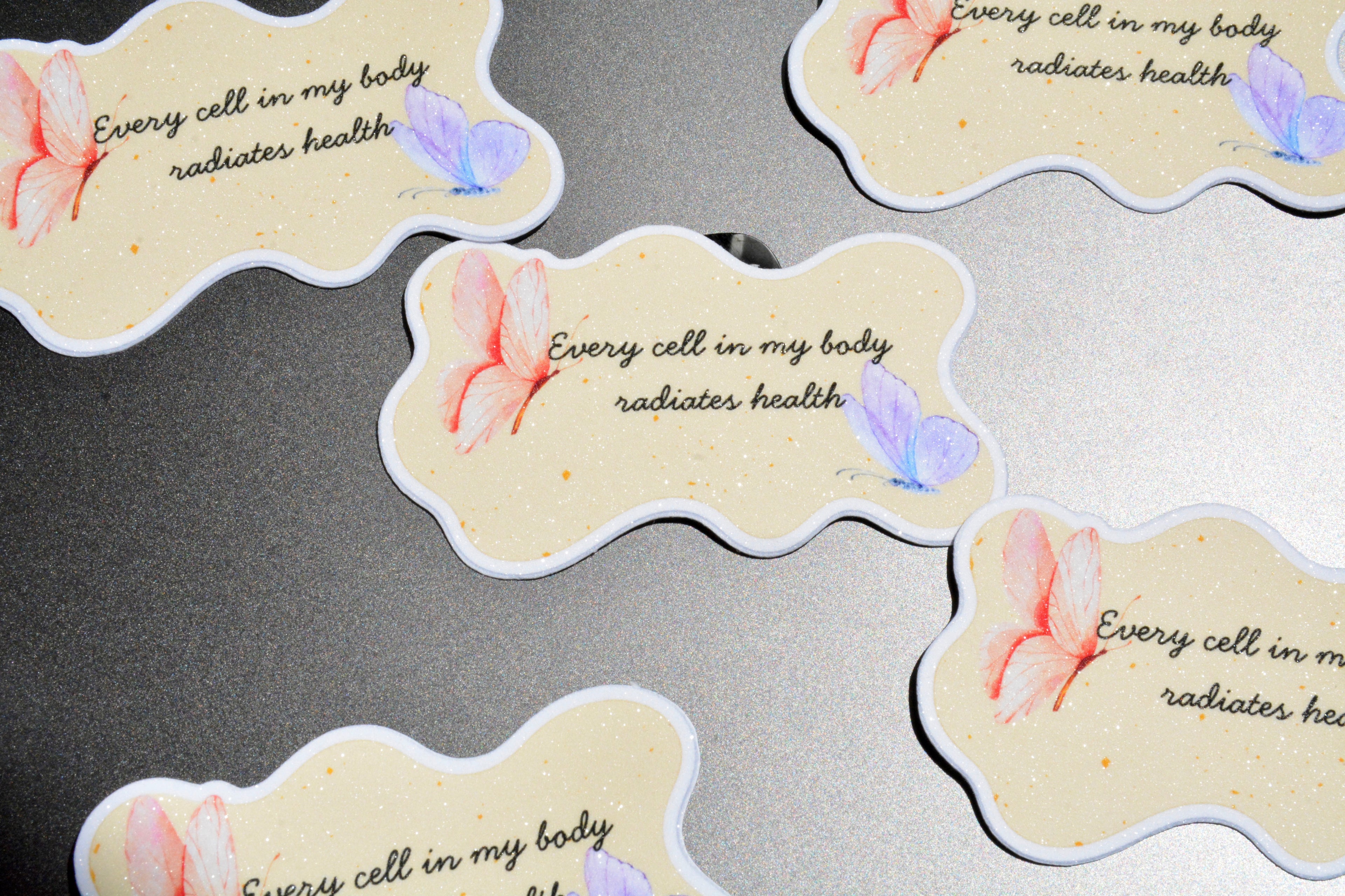 Several die-cut sticker tags with a wavy cloud shape are arranged on a gray textured surface. Each tag has a cream background with a small watercolor pink flower, a lavender butterfly, and the printed phrase 'Every cell in my body radiates health' in black script.