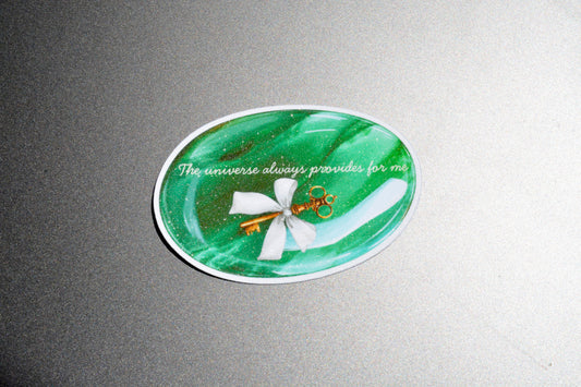 An oval green resin sticker with a white bow and gold key design, featuring the text 'The universe always provides for me' in white script, displayed on a gray textured background.
