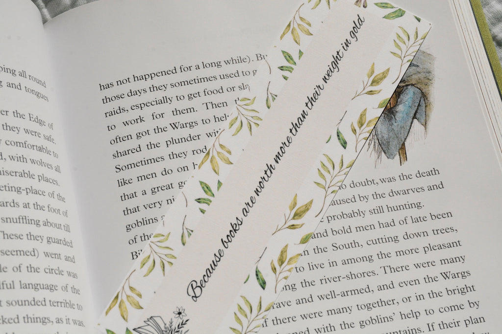 Square corner linen bookmark featuring the quote "Because books are worth more than their weight in gold" with watercolor greenery tucked inside an open fantasy book.
