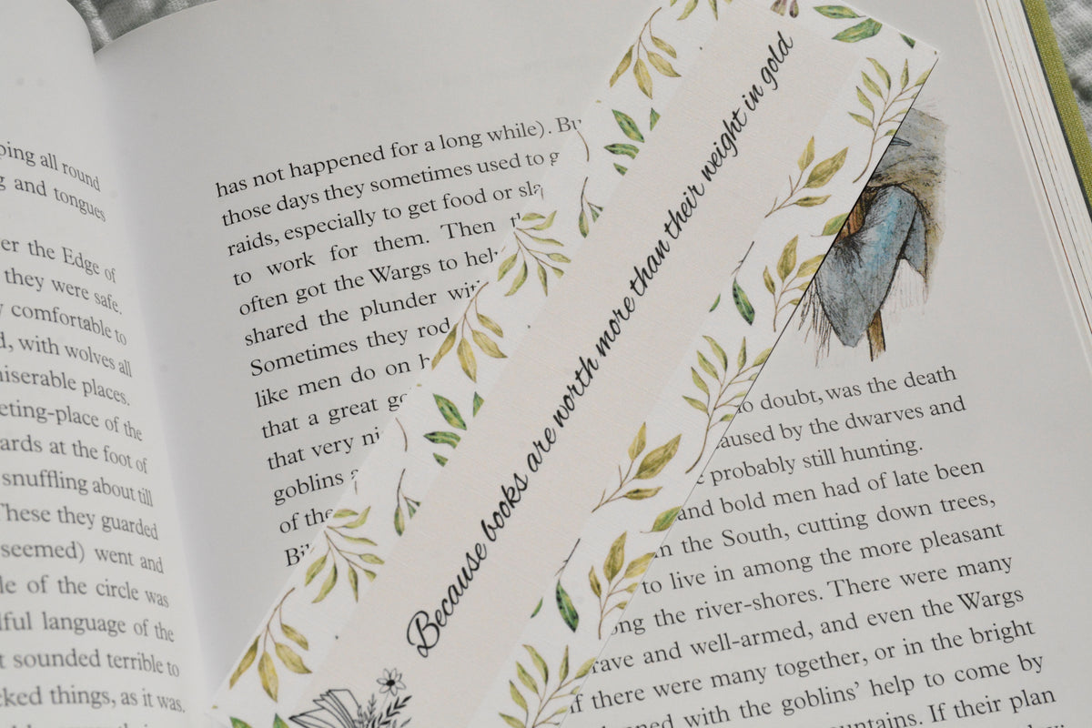 Square corner linen bookmark featuring the quote "Because books are worth more than their weight in gold" with watercolor greenery tucked inside an open fantasy book.