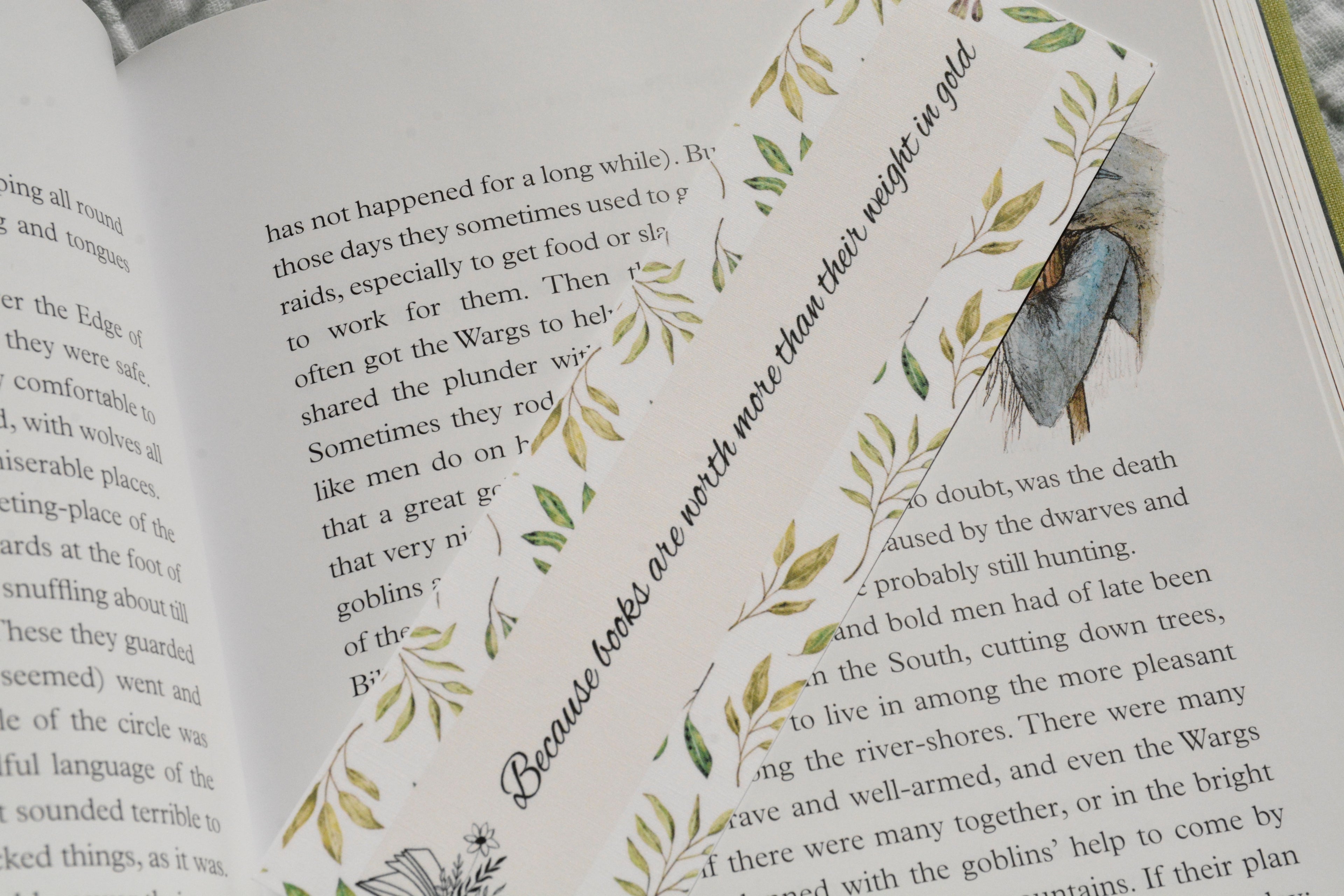 Square corner linen bookmark featuring the quote "Because books are worth more than their weight in gold" with watercolor greenery tucked inside an open fantasy book.