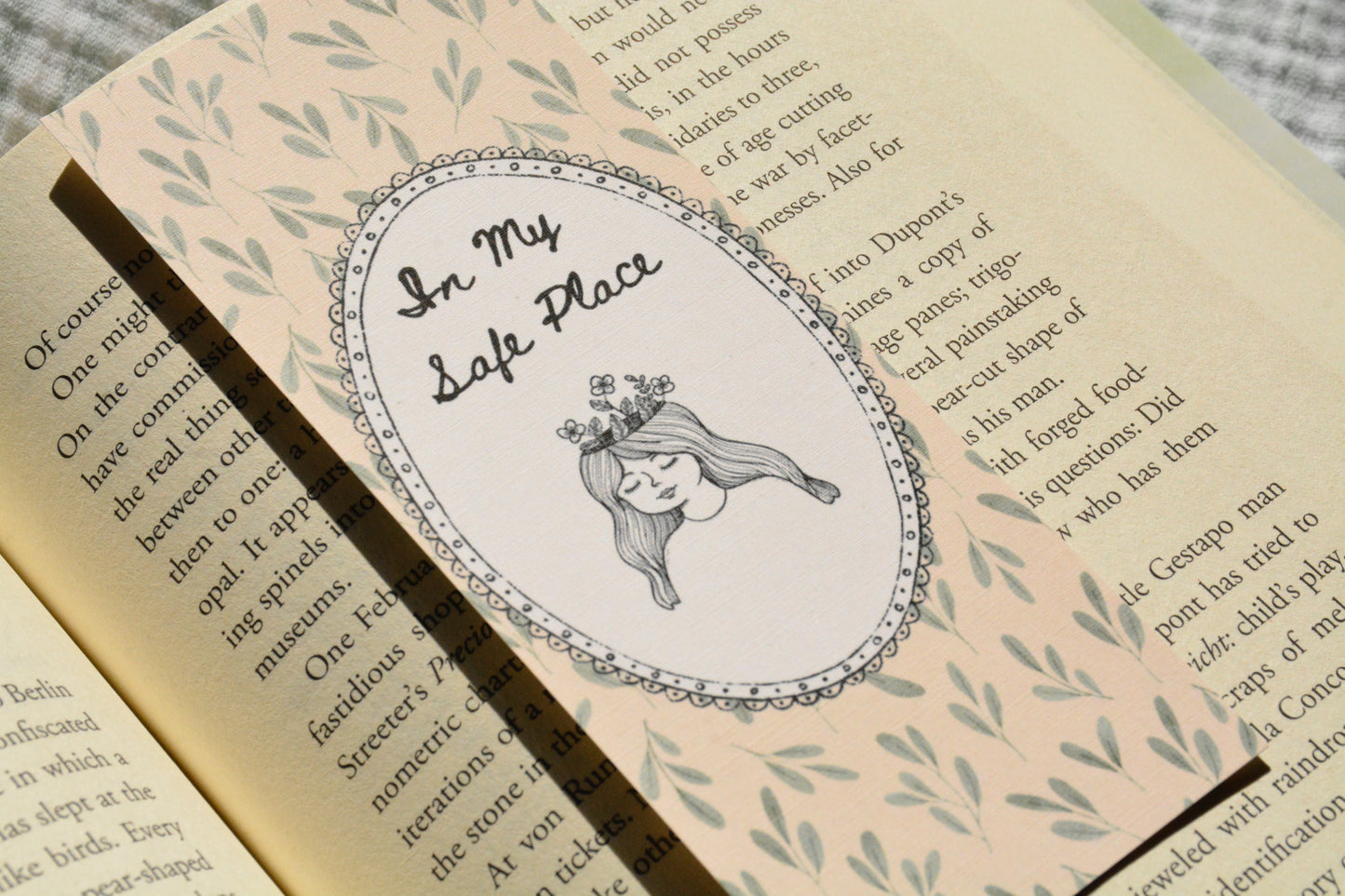 A close-up of a decorative book bookmark or sticker featuring an oval illustration with the text 'In My Safe Place' and a drawing of a girl with long hair, placed on an open book with printed text and leaf patterns.