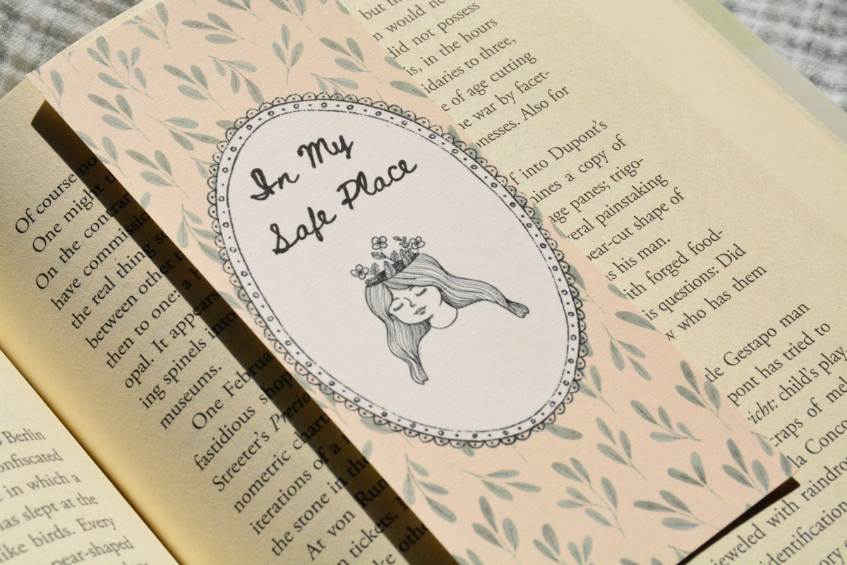 A close-up of a decorative book bookmark or sticker featuring an oval illustration with the text 'In My Safe Place' and a drawing of a girl with long hair, placed on an open book with printed text and leaf patterns.