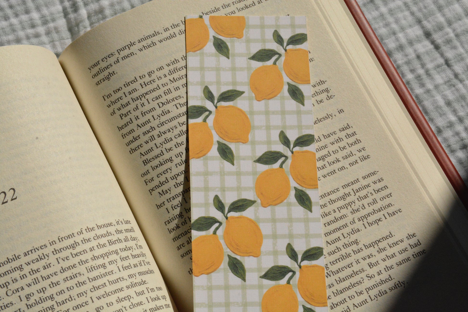 A rectangular book bookmark featuring a yellow lemon and green leaf pattern on a white gingham background, shown resting on the pages of an open book.